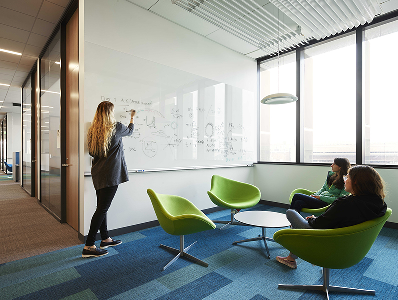 Graduate students sitting in soft green chairs with one student drawing on white board