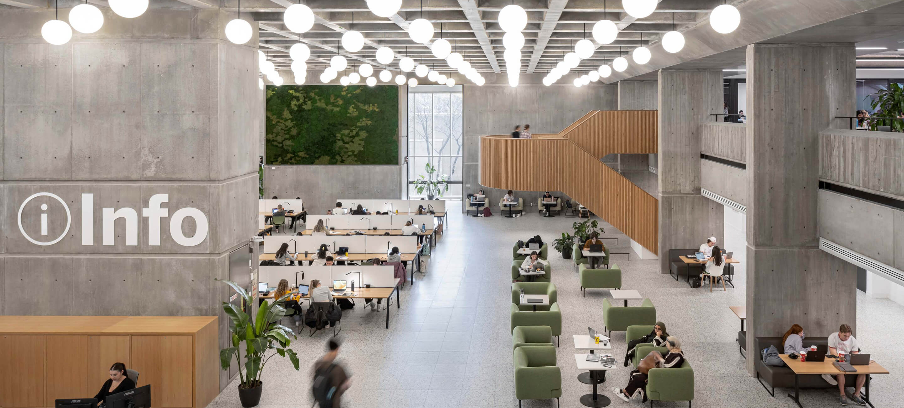 A view from the second story of a large room, with a circulation desk on the left side and wooden staircase on the right. Orb lights hang from the ceiling.